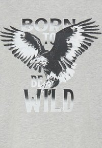 Camiseta gris con un gran gráfico en blanco y negro de un águila y el texto "NACIDO PARA SER SALVAJE" en una fuente en negrita en el centro.