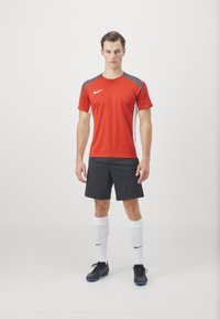 Červené športové tričko s šedými akcentmi, čierne šortky, biele ponožky a čierne futbalové topánky s modrými detailami. Materiál vyzerá ľahko a flexibilne.