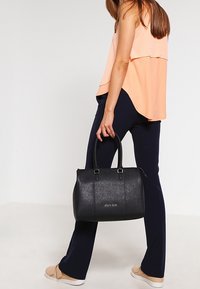 Kobieta w brzoskwiniowej bezrękawnikowej bluzce i granatowych spodniach trzyma czarną skórzaną torebkę, stojąc na tle jasnego gładkiego tła.