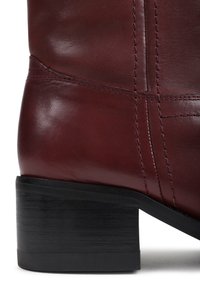 Bota de tornozelo de couro bordô com uma textura suave, featuring um salto bloco e sola preta em contraste. Detalhes de costura visíveis.