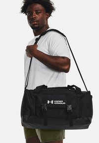 Bolsa de deporte negra con un fondo brillante, que cuenta con correas ajustables, múltiples compartimentos y el logo de Under Armour en blanco.