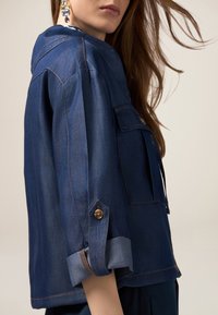 Chaqueta de denim azul oscuro con capucha, que presenta dos bolsillos frontales, mangas enrollables y adornos de botón marrón. Textura suave y diseño estructurado.