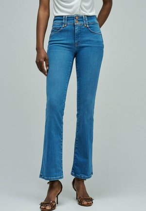 Personne portant un jean bleu taille haute avec fermeture à double bouton et des sandales à talons hauts marron à brides, debout devant un fond uni.