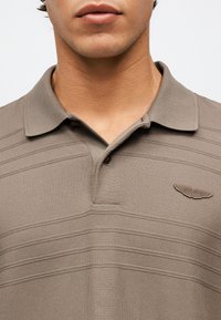 Άνδρας που φοράει πόλο πουκάμισο σε ταυπε απόχρωση με υφή, διπλωμένο γιακά και λογότυπο φτερών Aston Martin στο αριστερό στήθος.