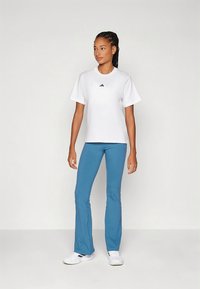 Λευκό t-shirt με κοντά μανίκια και μαύρο λογότυπο, σε συνδυασμό με μπλε φαρδιά παντελόνια και λευκά αθλητικά παπούτσια, τοποθετημένο σε απλό φόντο.