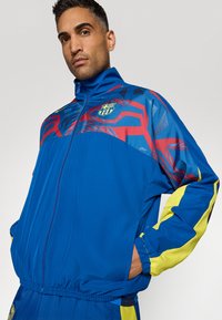 Modrá sportovní bunda s živými červenými a žlutými akcenty, zipem vpředu, vysokým límcem a logem na hrudi s grafickými vzory.