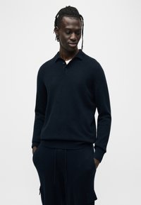 Πουλόβερ σε ναυτικό μπλε από απαλό πλεκτό ύφασμα με γιακά πόλο, τρία κουμπιά και ριμπ μανσέτες. Συνδυάζεται με αντίστοιχο σκούρο παντελόνι.