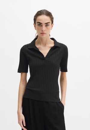 Jeune femme aux cheveux foncés portant un T-shirt noir en côte à manches courtes avec un col en V, debout devant un fond blanc uni.