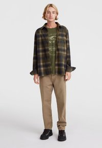 Flannel skjorte i grøn og marineblå tern, båret over en olivengrøn graphic T-shirt, kombineret med beige bukser og sorte snørebåndsstøvler.