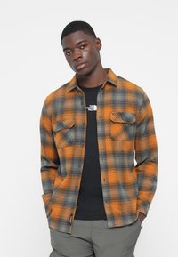 Camisa a cuadros naranja y gris con dos bolsillos en el pecho, frente abotonado y una camiseta negra de cuello redondo debajo. Tejido de mezcla de algodón.