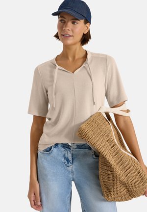 Femme portant un top beige à manches courtes, un jean bleu clair et une casquette bleue, tenant un grand sac en paille tressée avec des bretelles crème sur le bras.