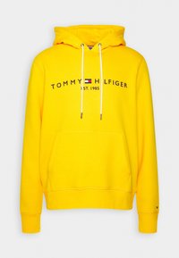Sudadera amarilla con un bolsillo frontal, cordones blancos y "TOMMY HILFIGER EST. 1985" impreso en negro en el pecho.