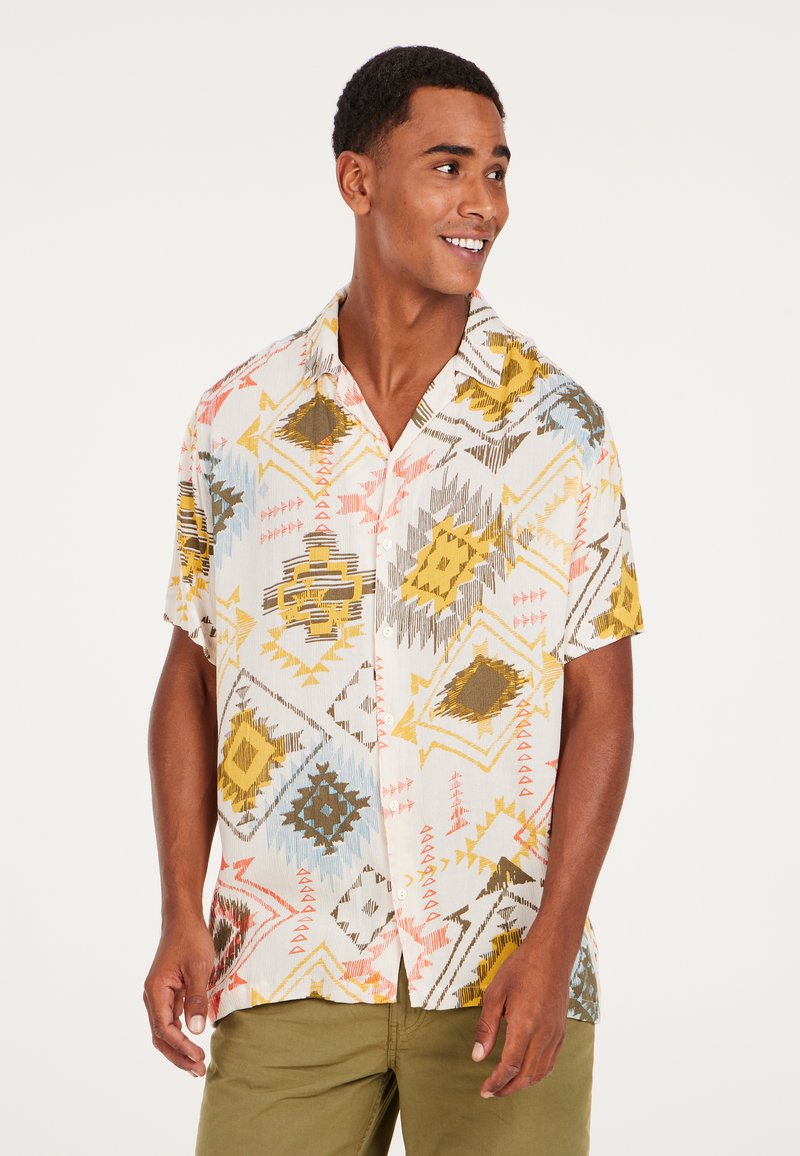 Camisa de botones de manga corta en tejido ligero, con coloridos patrones geométricos en amarillo, marrón, azul y rosa sobre un fondo blanco.