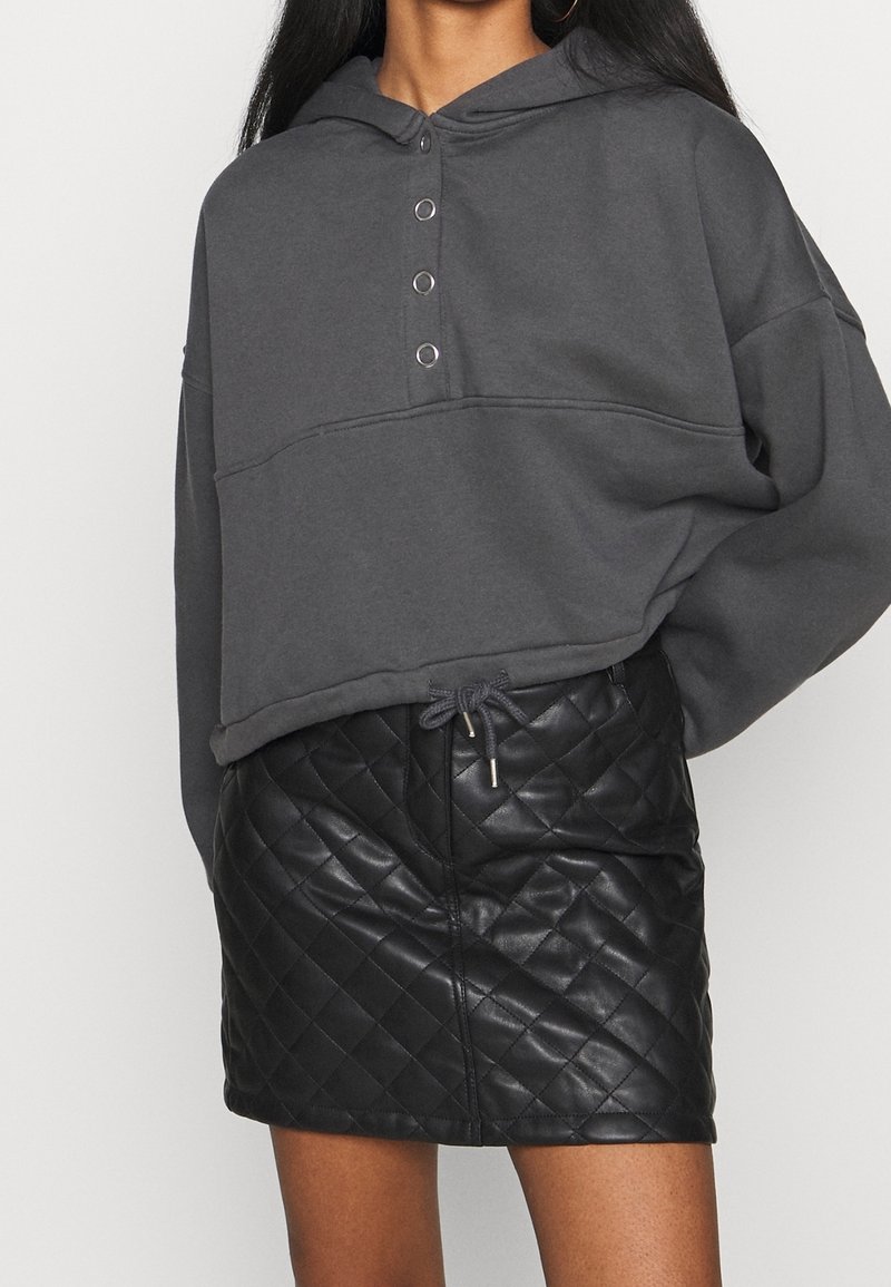 Femme portant un sweat à capuche court gris foncé avec des boutons en métal et une mini-jupe en cuir matelassé noire avec une taille à cordon.
