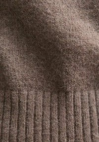Tejido de punto marrón con una superficie suave y texturizada que muestra un dobladillo acanalado. El material parece difuso con fibras finas en toda su extensión.