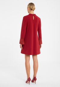 Rotes Kleid mit langen Ärmeln, Spitzenakzenten an den Manschetten und einem Schlüssellochverschluss im Rücken. Glatter Stoff, knielanges Design, kombiniert mit roten High Heels.