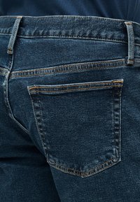 Gros plan sur la poche arrière d'un jean en denim bleu foncé avec des coutures orange visibles et une boucle de ceinture sur la taille.