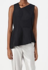 Top nero senza maniche con scollatura asimmetrica e orlo a peplo, realizzato in tessuto leggero; indossato con pantaloni argento lucidi.