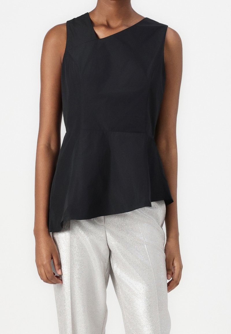 Top nero senza maniche con scollatura asimmetrica e orlo a peplo, realizzato in tessuto leggero; indossato con pantaloni argento lucidi.