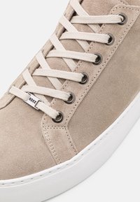 Baskets en daim beige avec des lacets blancs passés dans des œillets métalliques, dotées d'une étiquette en métal gravée "NBKK" sur une semelle en caoutchouc blanche.