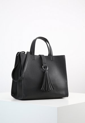 Sac à main en cuir noir texturé avec doubles poignées, une bandoulière et un grand pompon décoratif sur le devant, placé sur une surface blanche.