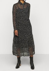 Svart genomskinlig klänning med vita spridda hjärttryck, långa ärmar, krage och en tärnad kjol, matchad med svarta klackade ankelkängor.