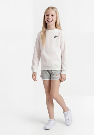 Ung pige med langt blondt hår smilende, iført hvid Nike sweatshirt, grå ellesse shorts og hvide sneakers, stående på hvid baggrund.