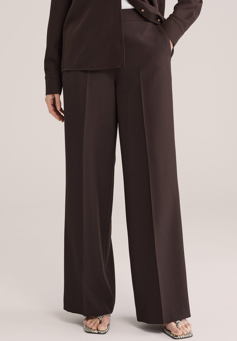 Vrouw draagt donkerbruine brede pijpen broek met plooi aan de voorkant en open teen sandalen met slangenhuidprint, staand met een hand in de zak.