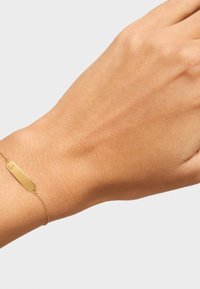 Pulsera de oro con una etiqueta rectangular, cadena fina y textura suave, llevada en una mano sobre un fondo neutro.