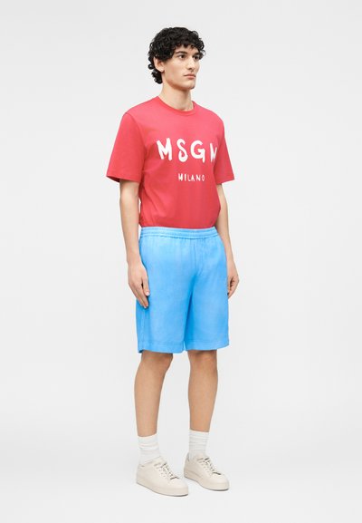 Νεαρός άνδρας στέκεται, φοράει κόκκινη μπλούζα MSGM Milano, ανοιχτό μπλε σορτς, άσπρες κάλτσες και μπεζ αθλητικά παπούτσια μπροστά σε απλό γκρι φόντο.