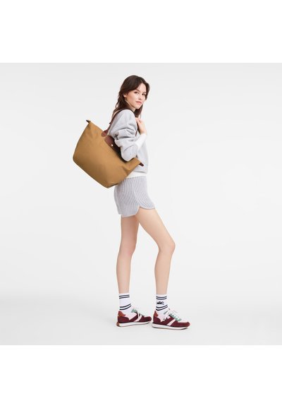 Braune Tragetasche mit glatter Textur und Lederakzenten, getragen über einem grauen Pullover und gerippten Shorts, kombiniert mit mehrfarbigen Sneakers.