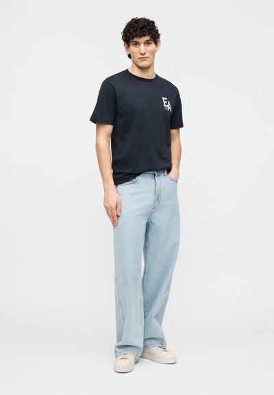 Άνδρας με σγουρά μαλλιά στέκεται αντιμετωπίζοντας προς τα εμπρός φορώντας μαύρο μπλουζάκι με λογότυπο «EA», ανοιχτό μπλε τζιν παντελόνι με φαρδιά πόδια και μπεζ αθλητικά παπούτσια.