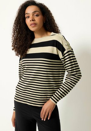 Woman with curly hair wearing a beige and black striped long-sleeve sweater with buttons on the shoulder and black pants against a white background.