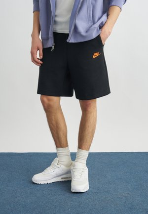 Személy fekete Nike rövidnadrágot visel narancssárga logóval, fehér zoknival, fehér Nike sportcipővel, valamint világos lila, cipzáros felsőt egy fehér póló felett.