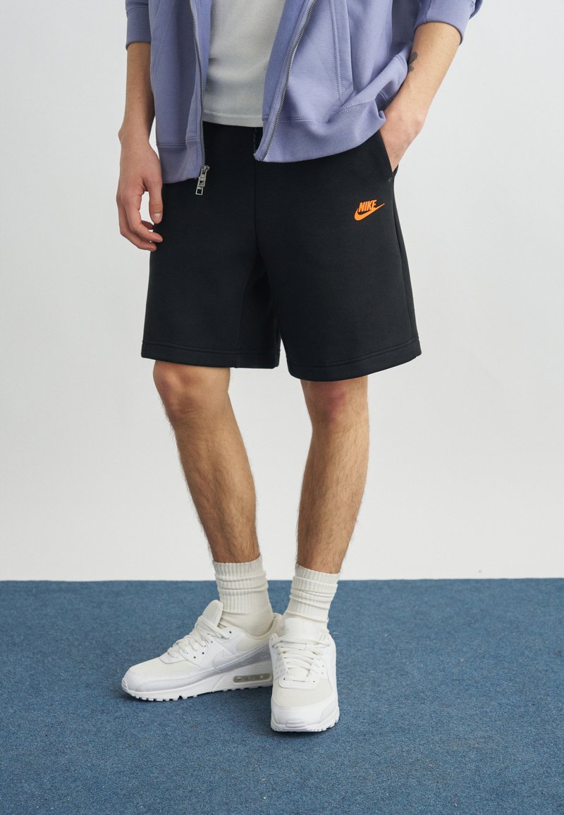 Άνθρωπος που φοράει μαύρα σορτς Nike με πορτοκαλί λογότυπο, λευκές κάλτσες, λευκά αθλητικά παπούτσια Nike και μωβ ανοιχτό μπουφάν με φερμουάρ πάνω από λευκό πουκάμισο.