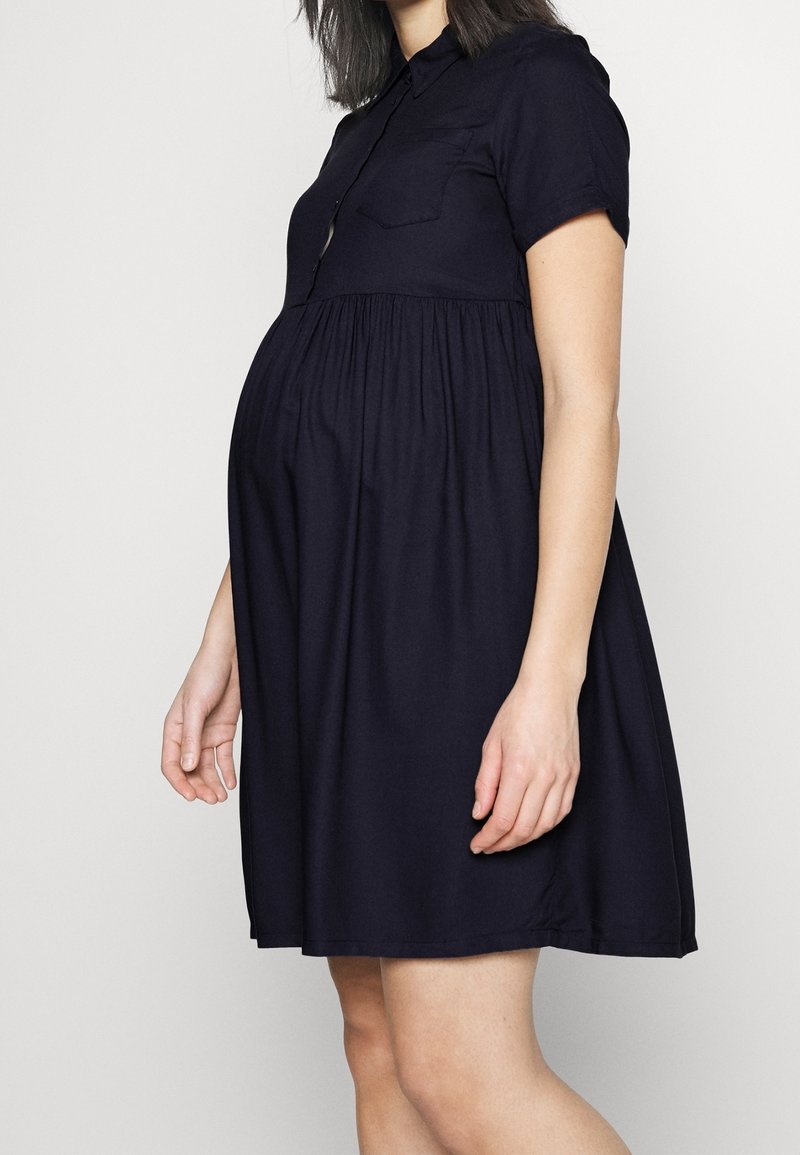 Femme enceinte portant une robe bleu marine à manches courtes, longueur genoux, avec un col et une poche poitrine, debout devant un fond uni.