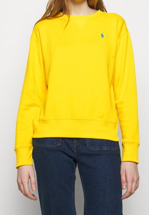 Personne portant un sweat-shirt jaune vif à manches longues avec un petit logo bleu sur la poitrine gauche et un jean bleu foncé.