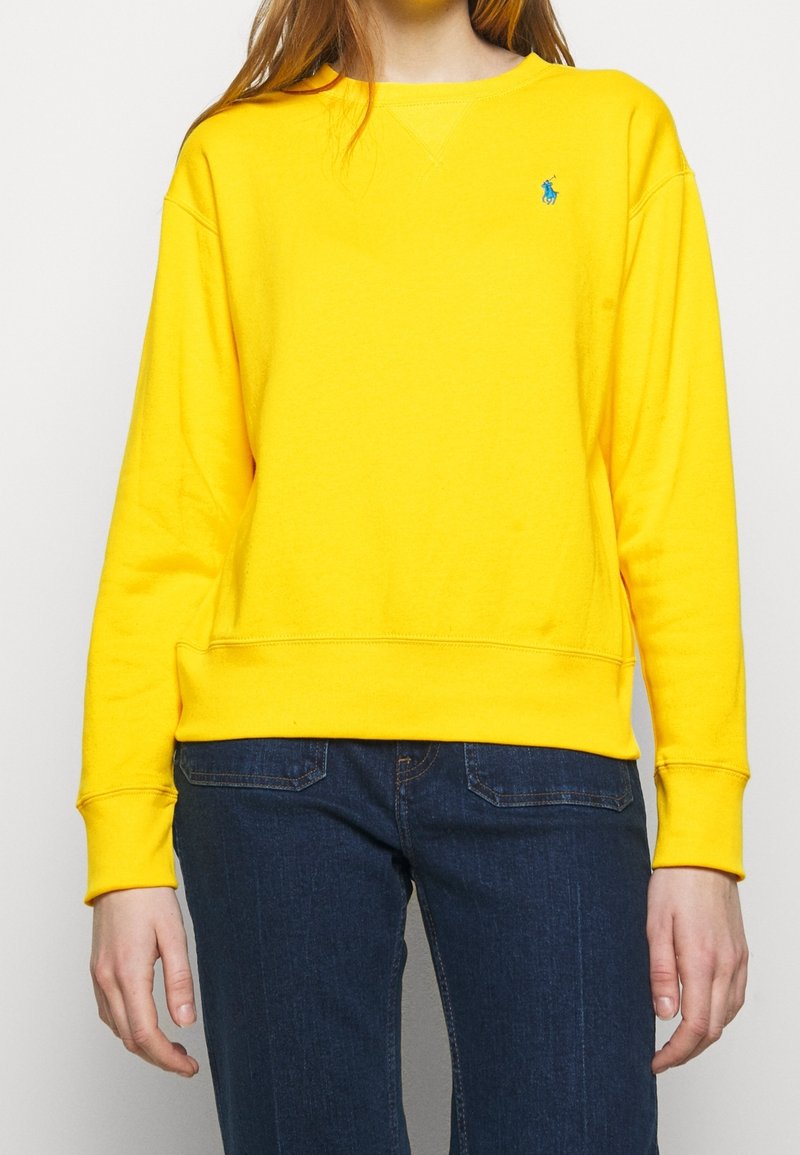 Personne portant un sweat-shirt jaune vif à manches longues avec un petit logo bleu sur la poitrine gauche et un jean bleu foncé.