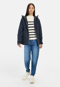 Marineblauwe waterdichte jas met een capuchon, ritssluiting en elastische manchetten, gedragen over een crèmekleurige en marineblauwe gestreepte trui en lichtblauwe spijkerbroek.