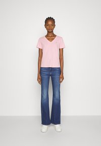 Camiseta de algodón suave de cuello en V en color rosa, combinada con jeans acampanados de un azul oscuro. La modelo lleva zapatillas blancas. Diseño simple con una marca mínima.