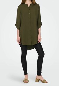 Olivgrünes langes Hemd mit durchgehendem Knopfverschluss, dreiviertel Ärmeln und abgerundetem Saum, kombiniert mit schwarzen Leggings und olivgrünen Sandaletten mit Absatz.