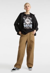 Giacca leggera nera sopra una maglietta nera con grafica a design floreale, abbinata a pantaloni beige larghi e sneaker nere con dettagli bianchi.