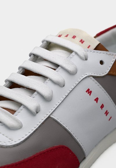 Baskets en cuir blanc et gris avec des accents en daim rouge, dotées de lacets plats blancs, d'une languette à logo, et d'une finition lisse et texturée.