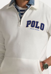 Man med vit zip-sweatshirt med blå "POLO Ralph Lauren" logotyp, blå chambray-skjorta, randig t-shirt och grön klocka med brun rem.