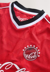 Maglia sportiva rossa con collo a V nero e bianco con logo Coca-Cola e trama del tessuto a motivo.