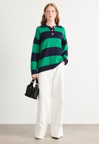 Grün- und navygestreifter gestrickter Pullover mit Kragen und drei Knöpfen, kombiniert mit weißen weit geschnittenen Hosen und einer kleinen schwarzen Handtasche.