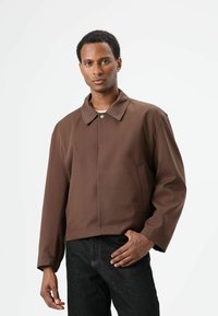 Lichtgewicht bruine jas met een puntige kraag, knoopsluiting, zijzakken en een ontspannen pasvorm, gedragen over een donkere jeans. Gladde textuur.