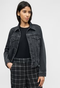 Γυναίκα με μακριά σκούρα μαλλιά που φοράει μαύρο τζιν μπουφάν με καρφιά, μαύρο τοπ και μαύρο-άσπρο καρό παντελόνι, με το ένα χέρι στην τσέπη.