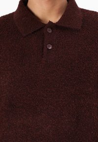 Polo in pile color bordeaux con superficie strutturata, chiusura con bottoni e design del colletto classico. Bottoni lisci aggiungono un dettaglio sottile.