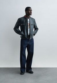 Bărbat stă cu mâinile în buzunare, purtând o jachetă de piele neagră, un tricou alb cu imprimeu, blugi albăstrui închis și adidași negri, pe un fundal simplu.